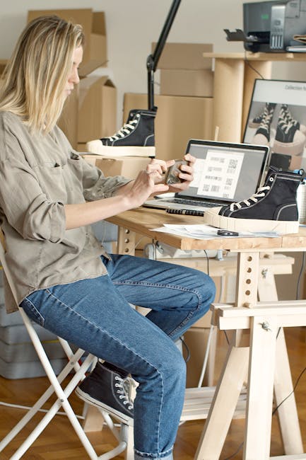 Woman using laptop and smartphone to manage an online shoe store in a workspace filled with boxes.